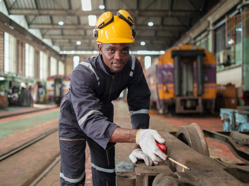 A focused industrial technician in a yellow hard hat uses a laptop while standing next to heavy machinery in a factory setting.