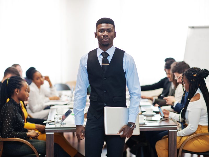 Face of handsome african business man, holding laptop on the background of business peoples multiracial team meeting, sitting in office table.